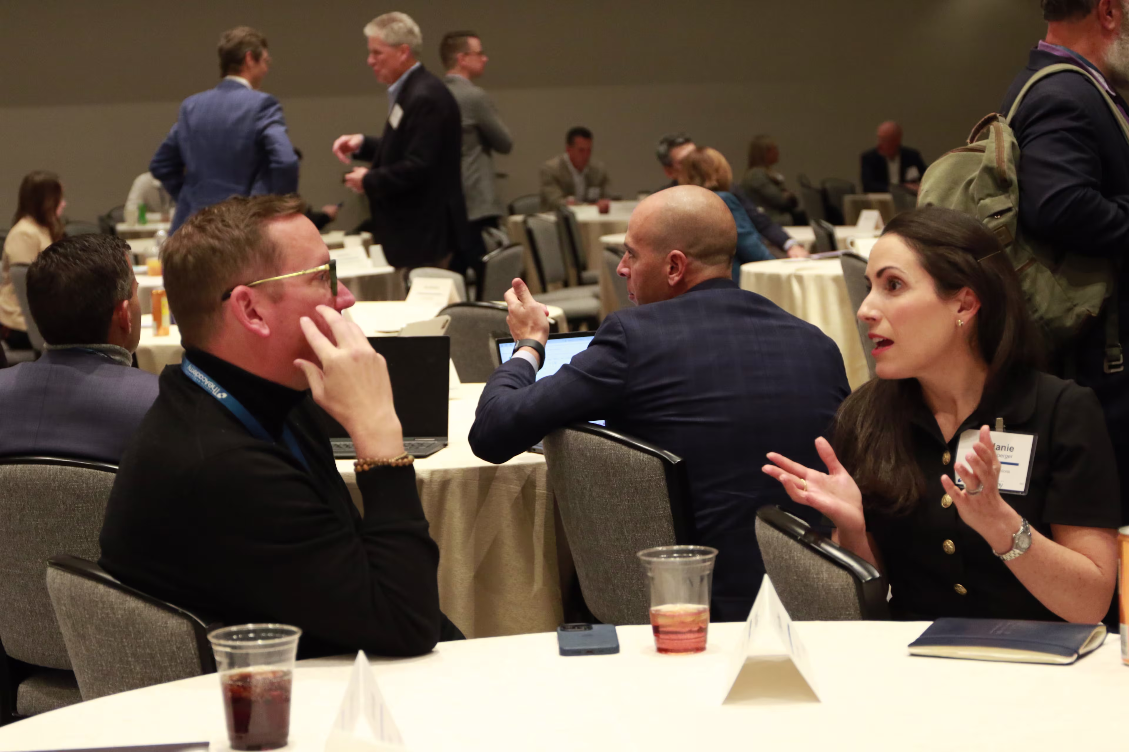Two attendees engage in an animated conversation at a roundtable during a professional event. Other participants are seated and conversing at nearby tables in a large, well-lit conference room. Notepads, drinks, and name cards are visible on the tables, reflecting a collaborative and interactive setting.

