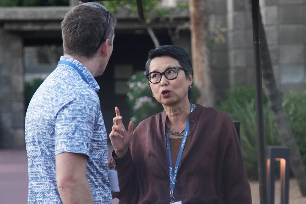 A woman gestures while speaking with a man during an outdoor conversation at a professional event. Both are wearing conference badges and appear engaged in discussion.