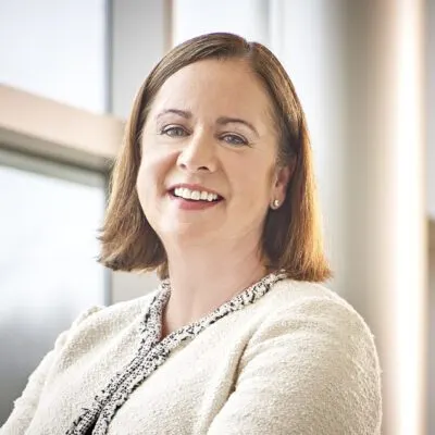 Woman with shoulder-length brown hair wearing a cream-colored textured jacket, smiling while seated near a large window with soft natural lighting.