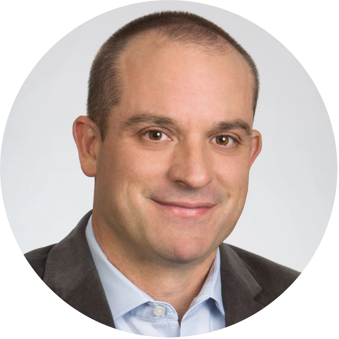 Man with close-cropped hair wearing a gray blazer and light blue button-up shirt, smiling against a neutral light gray background.