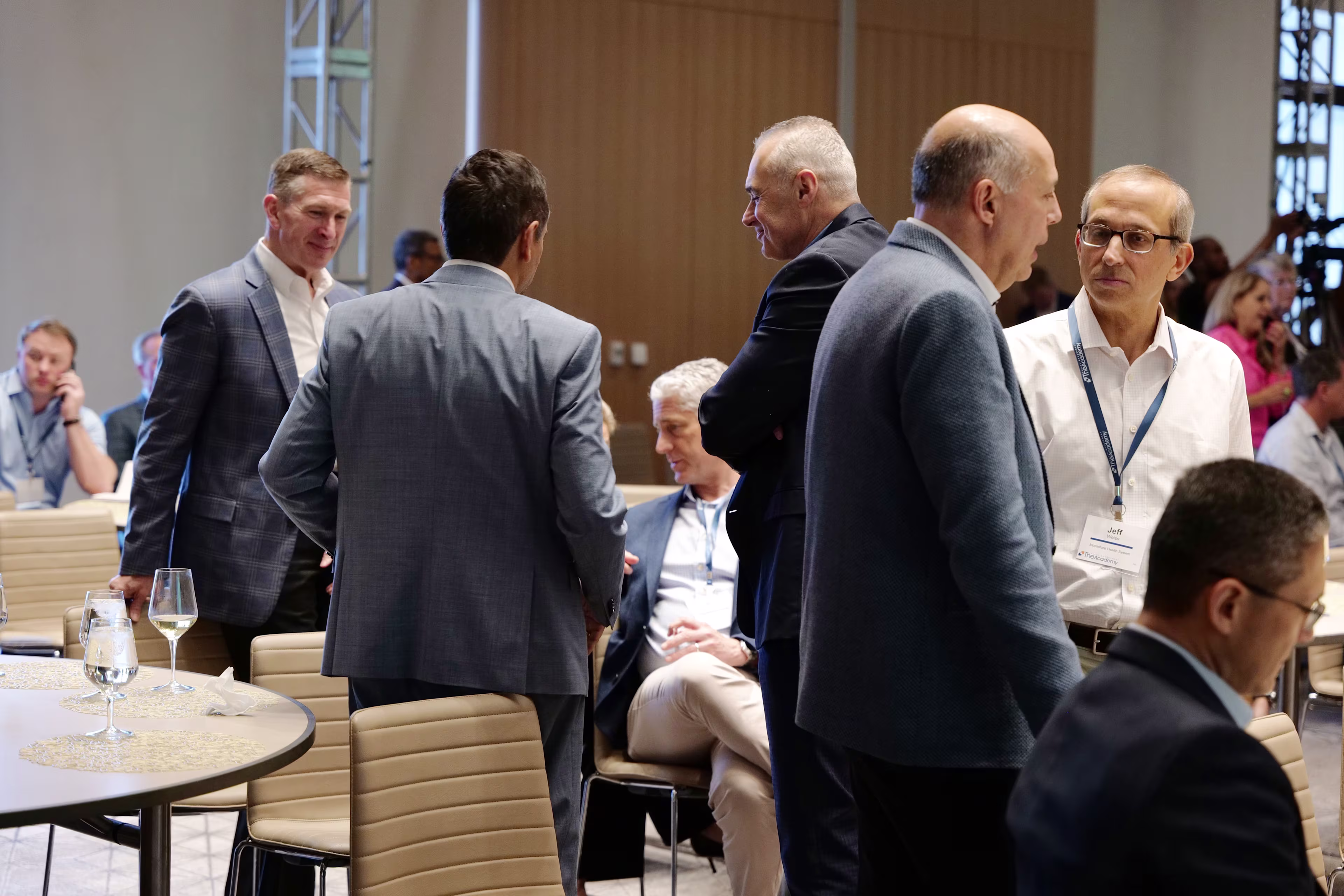 Groups of professionally dressed attendees are seen conversing and networking in a large, modern conference room. Several people are standing and engaging in discussion, while others are seated at round tables. The atmosphere is professional and collaborative, with natural light filtering in from large windows.

