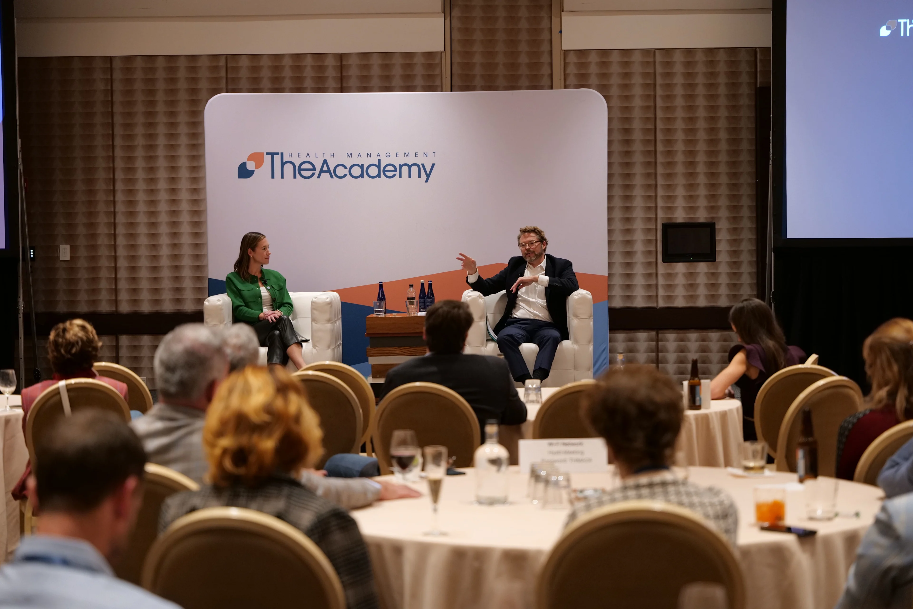 Conference session with two speakers seated on stage in white chairs, engaged in discussion. A woman in a green blazer and a man in a dark jacket speak in front of a backdrop displaying The Health Management Academy logo. Audience members sit at round tables facing the stage.
