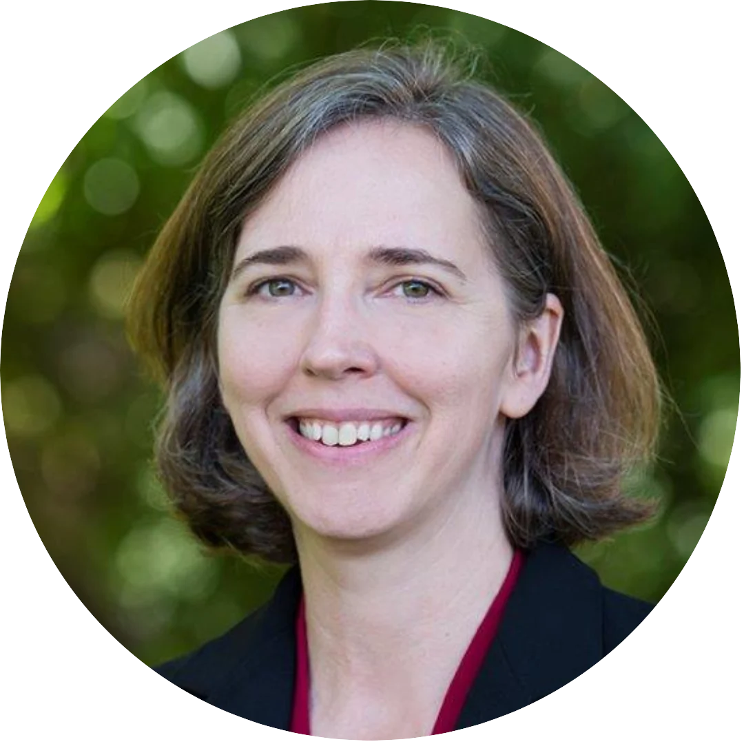 Smiling woman with short brown hair wearing a dark blazer and red top, photographed outdoors in front of a blurred green leafy background.
