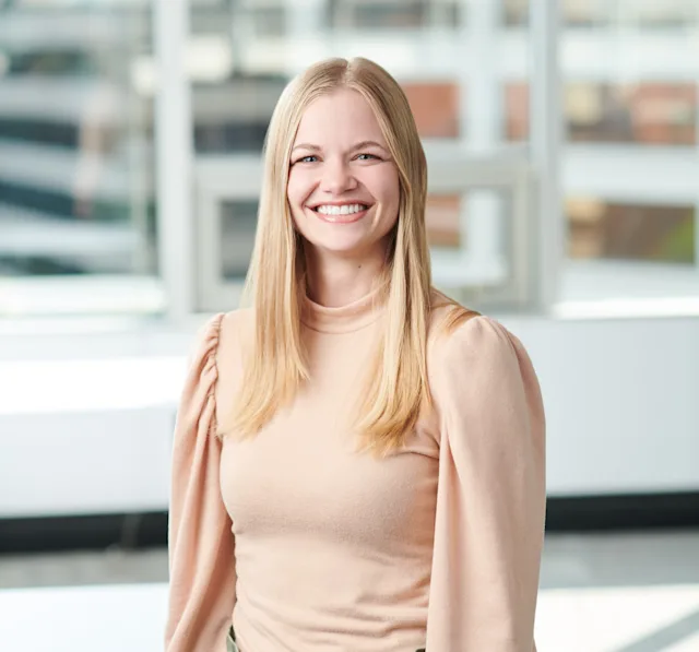 A woman with long straight blonde hair smiles brightly while wearing a light pink long-sleeve top with puffed shoulders. She stands in front of large windows with a modern office building in the background.