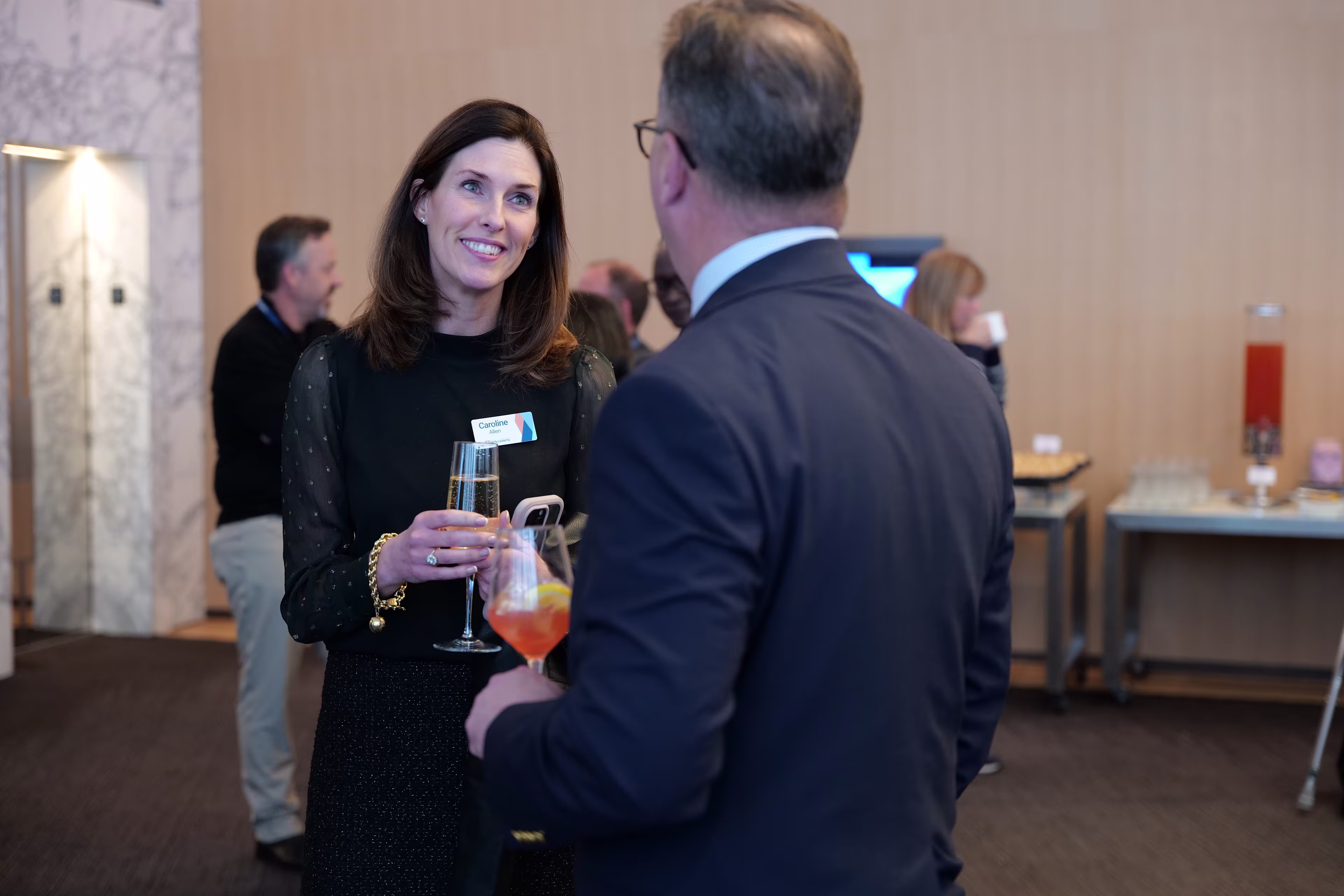 A woman and a man engage in conversation while holding drinks during a networking event, with other attendees and refreshments visible in the background.
