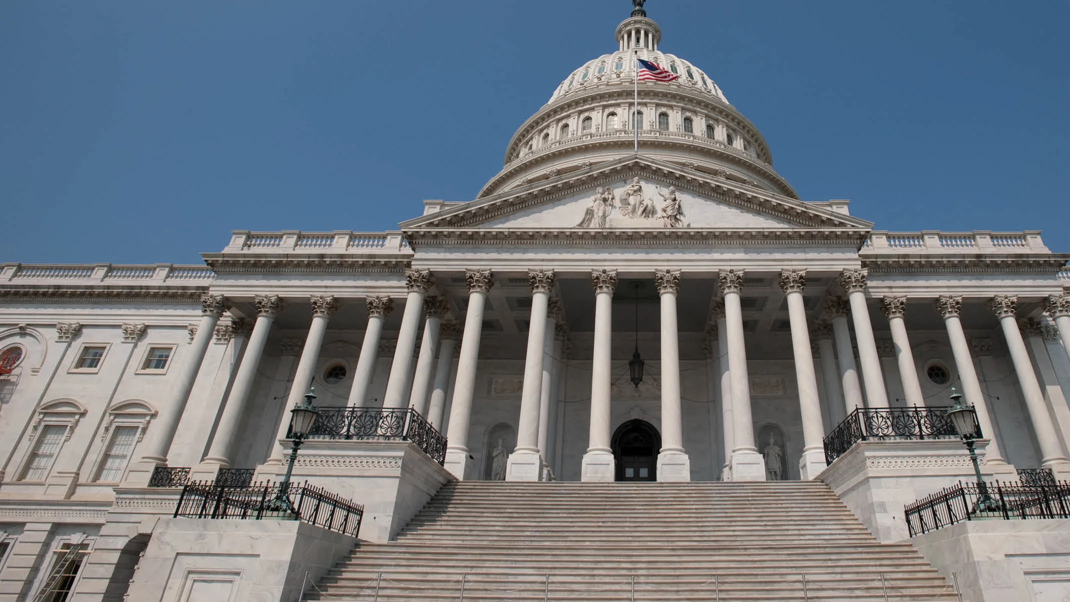 View of the U.S. Capitol Building’s east front with a clear blue sky in the background, showcasing the iconic dome, Corinthian columns, and grand staircase. 