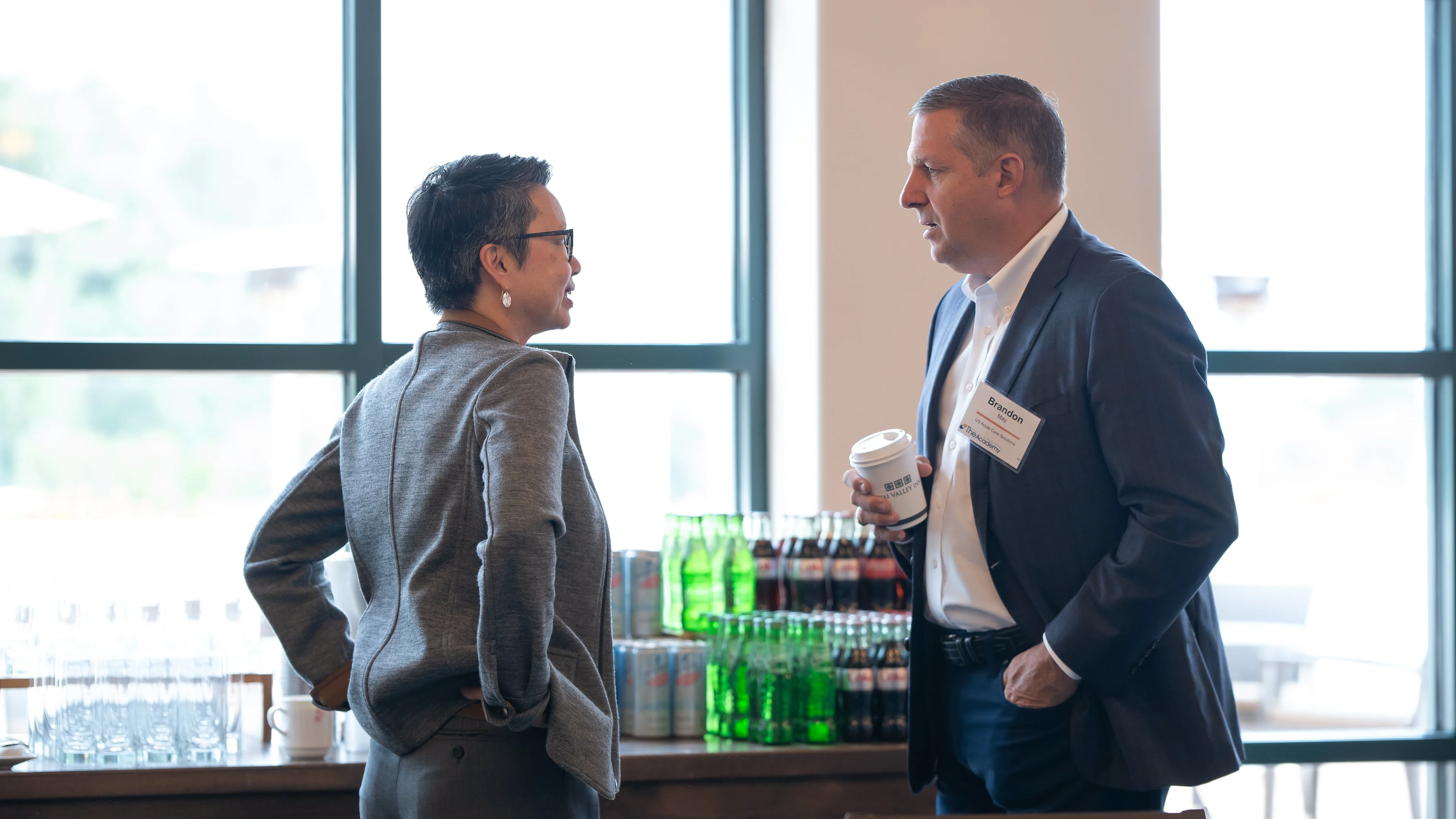 Two conference attendees stand and talk near a beverage station stocked with bottled drinks, with natural light streaming in from large windows behind them.