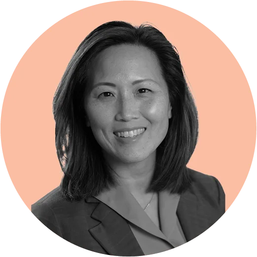 Black and white image of a woman with shoulder-length hair wearing a blazer and blouse, smiling against a circular peach-colored background.