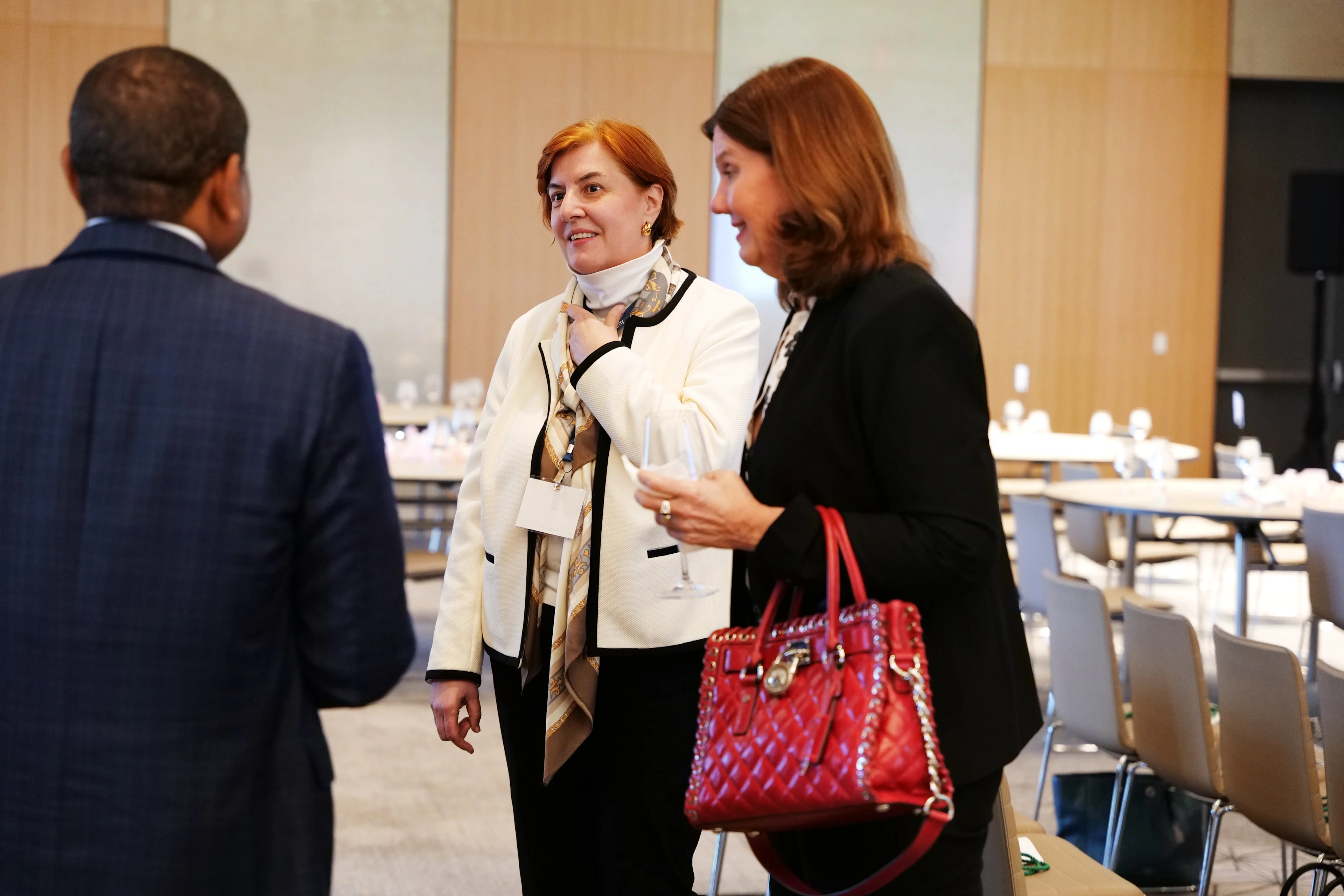 Two women and one man are engaged in a conversation inside a large conference room. One woman wears a white blazer and scarf, while the other carries a red handbag. They appear to be networking between sessions at a healthcare leadership event, with rows of chairs and tables in the background.

