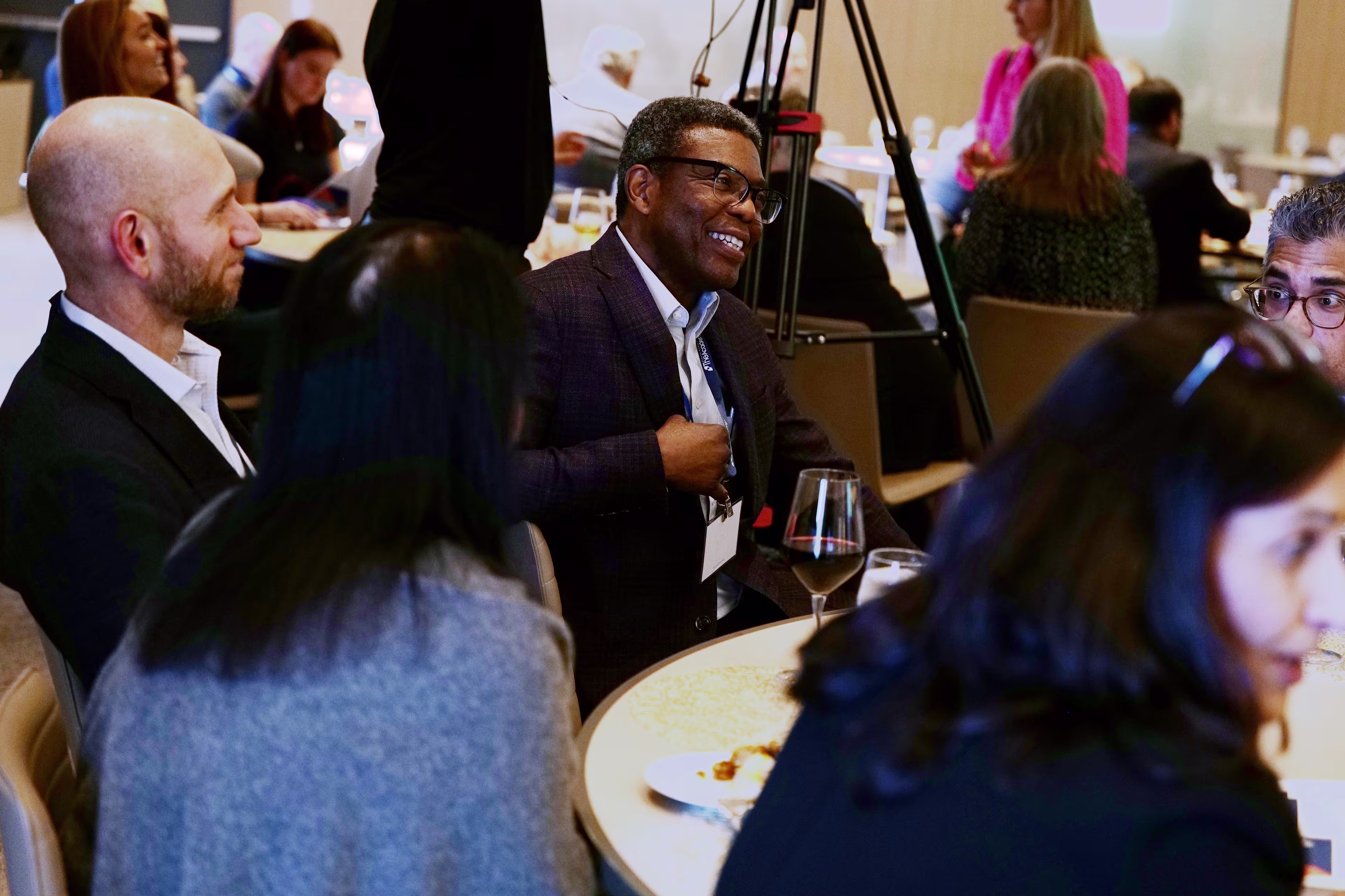 Attendees sit around a round table engaged in conversation during a professional event. A man in a suit is smiling and speaking, while others listen attentively. Plates, glasses, and a wine glass suggest a meal is being shared. In the background, additional participants and a camera tripod are visible, indicating a formal yet relaxed setting.

