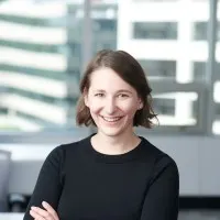  A woman with short brown hair wearing a black top smiles with her arms crossed, standing in a modern office space with large windows in the background. 