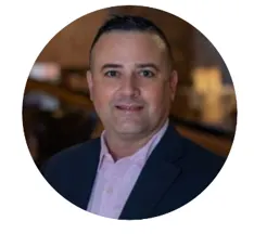 A man with short dark hair wearing a dark blazer and light pink shirt smiles in a professional headshot taken in a warmly lit indoor setting.
