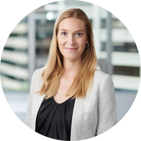 A woman with long blonde hair, wearing a light blazer and black top, poses for a professional headshot in front of an office window backdrop.