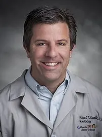 A smiling man in a light gray lab coat poses for a professional headshot against a neutral studio background.