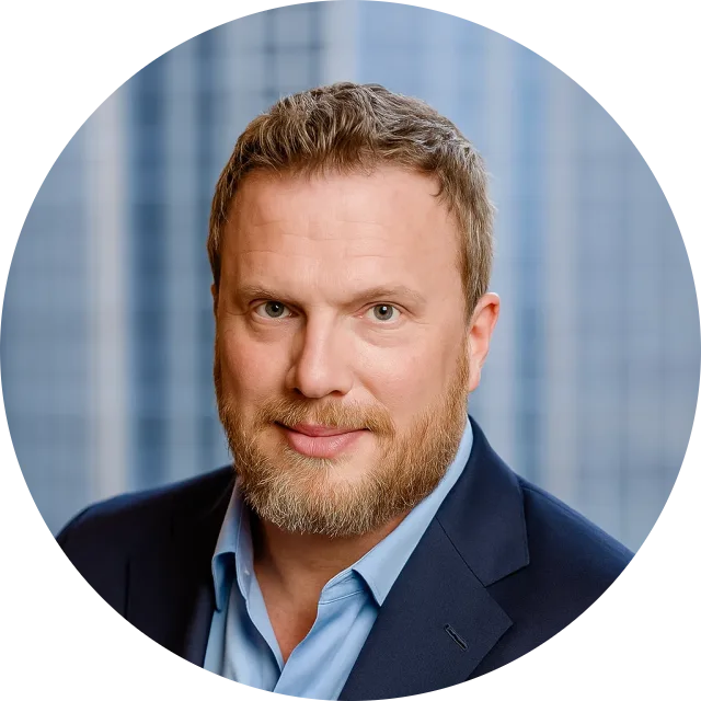 A man with short blonde hair and blonde beard, wearing a dar blue suit and a light blue button up, poses for a professional headshot with blue curtains in the backdrop.