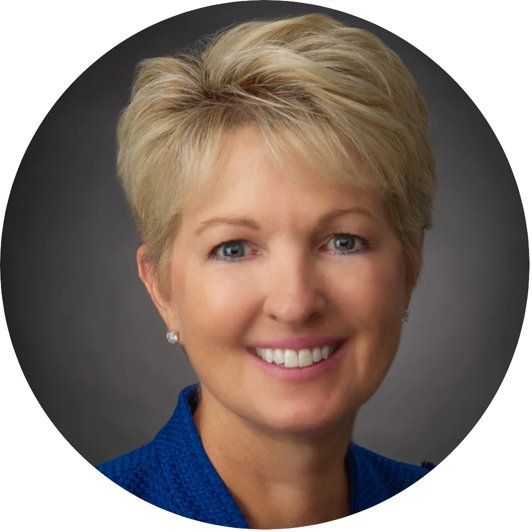Smiling woman with short blonde hair wearing a royal blue blazer and stud earrings, posed against a dark gray studio background.