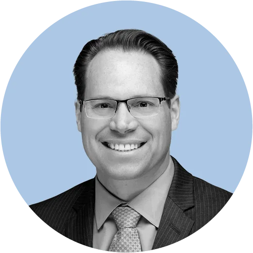 A smiling man in glasses and a suit with a tie is photographed in a grayscale image over a circular light blue background.