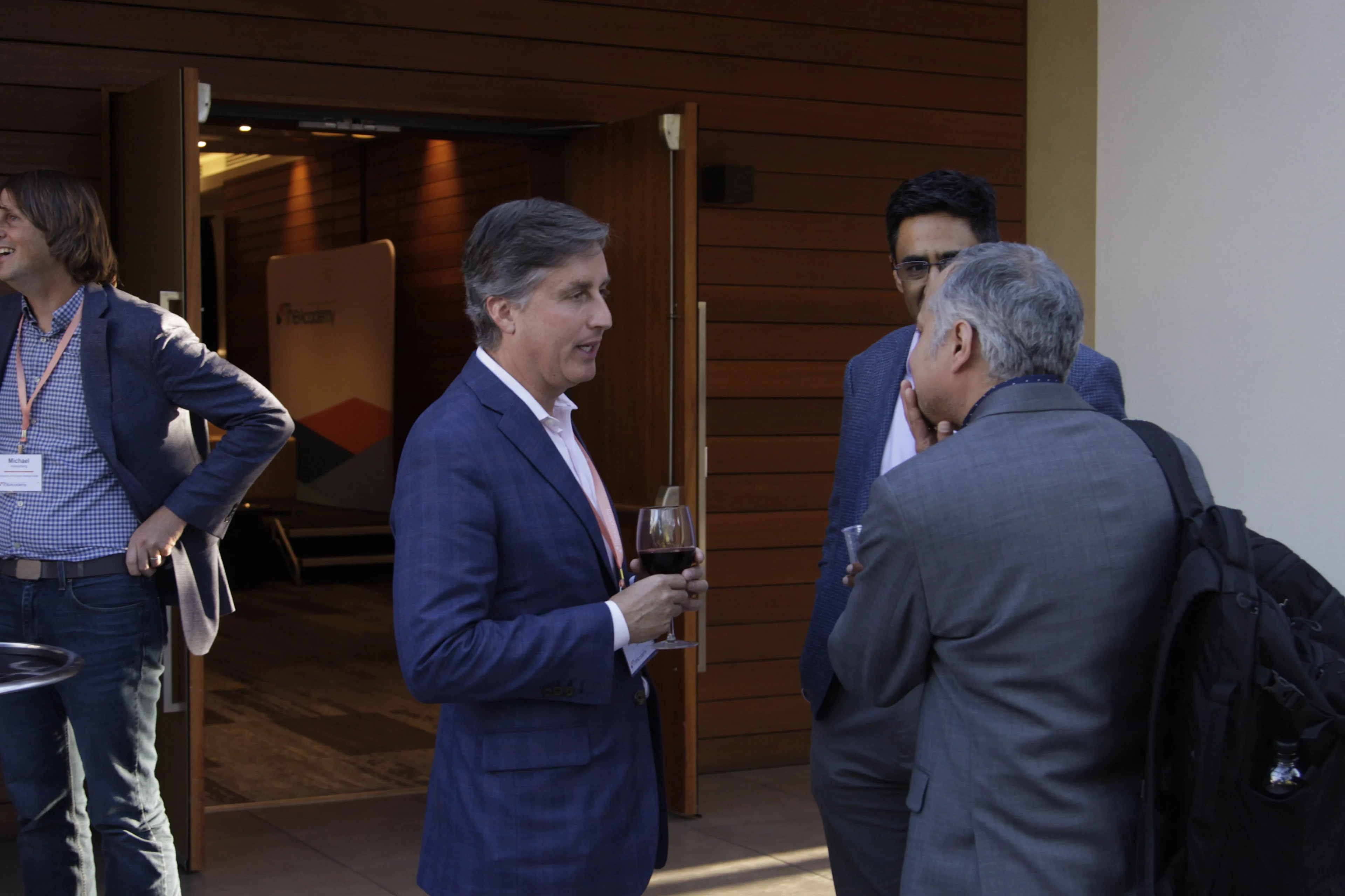 Several professionally dressed attendees are seen talking outside a conference room. One man holds a glass of wine while engaging in conversation with two others. The open doors behind them reveal an event space with branded signage. The setting suggests a casual networking moment during a break in programming.

