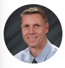 A man with short-cropped brown hair wearing a light blue dress shirt and patterned tie smiles for a professional headshot against a dark, softly lit background.