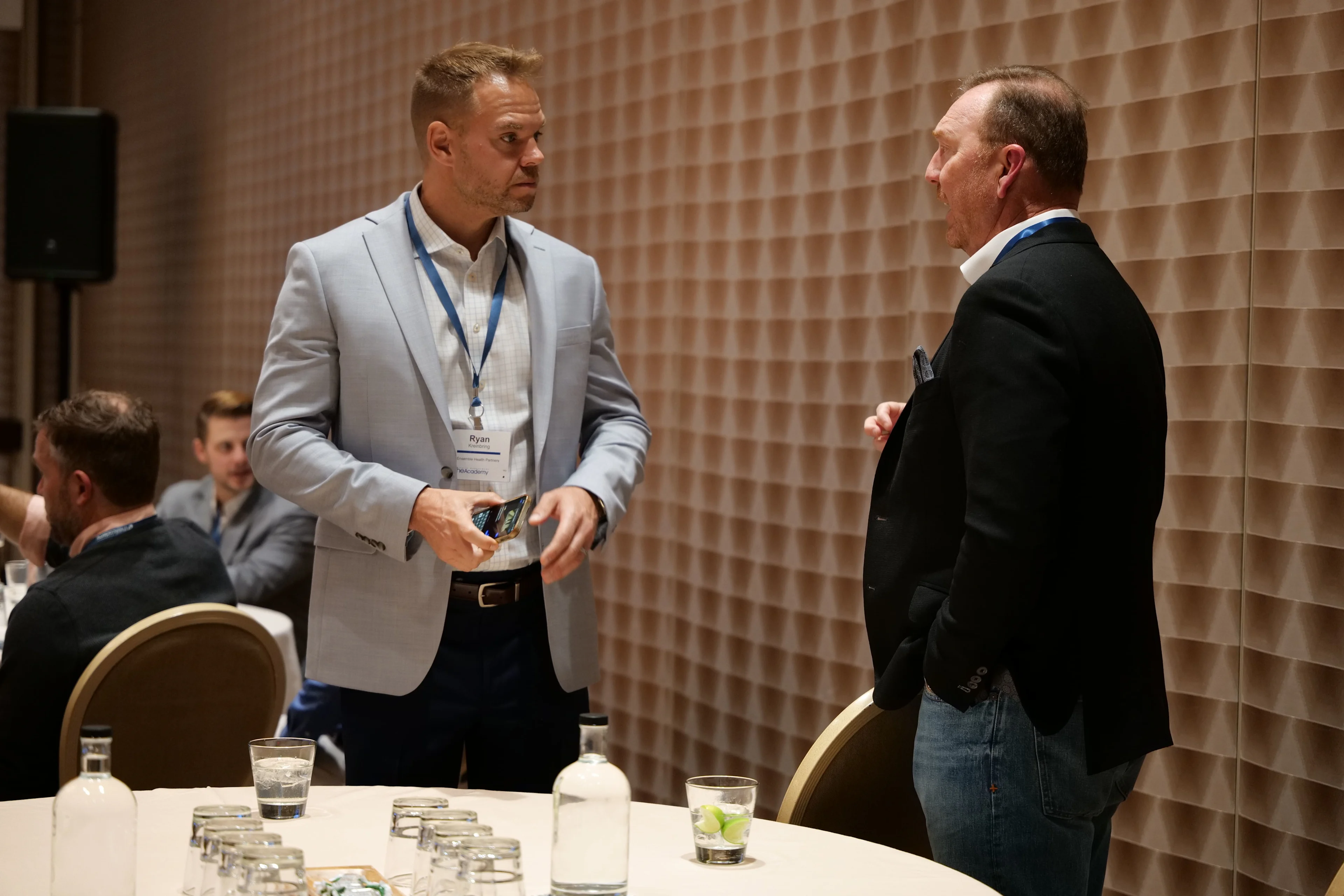 Two men stand and talk during a conference session. The man on the left, wearing a light gray blazer and conference badge labeled "Ryan," holds a snack packet while listening to the man on the right, who is dressed in a black blazer and jeans. In the foreground, a round table is set with bottled water, drinking glasses, and lime wedges. Other attendees are seated and conversing in the background.

