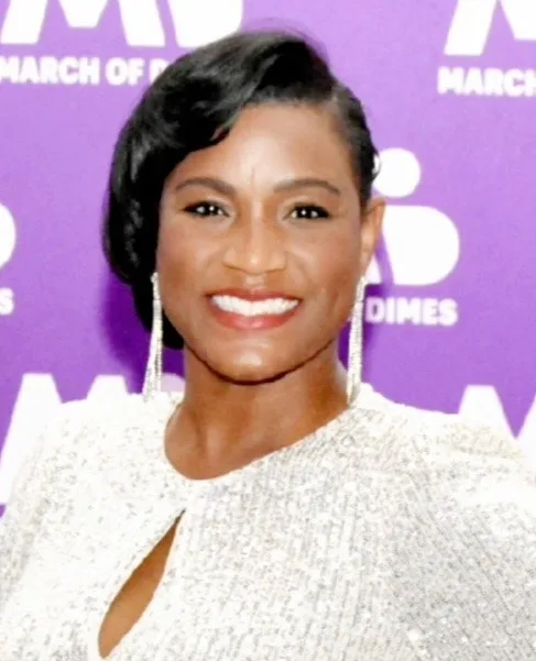 Smiling woman in a sparkly silver outfit with side-swept hair and long earrings, standing in front of a purple March of Dimes event backdrop.