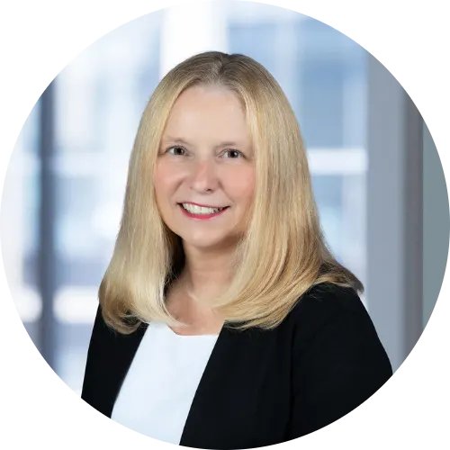 Professional headshot of a smiling woman with straight, shoulder-length blonde hair, wearing a black blazer over a white blouse, standing in front of a blurred office background with large windows.