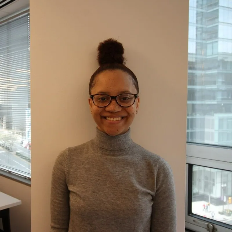 Smiling woman wearing glasses and a gray turtleneck sweater, with her hair in a high bun, standing in front of a white wall near large office windows overlooking a city street.