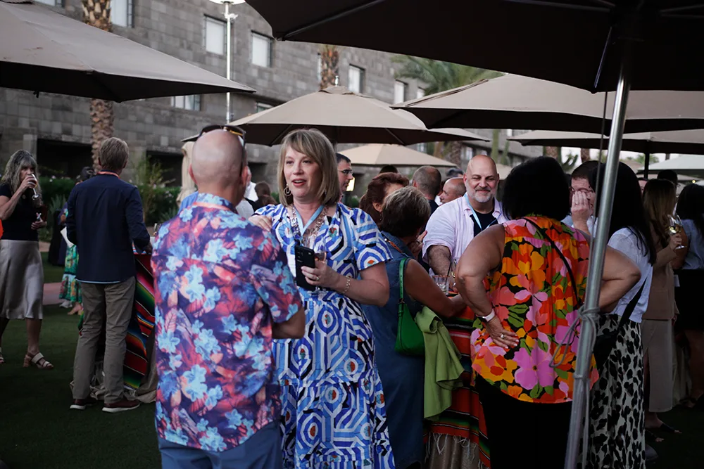 Attendees gather under patio umbrellas at an outdoor networking event, engaging in conversation while dressed in colorful, casual attire.