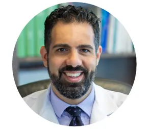 A smiling man with dark hair and beard wearing a white medical coat and blue dress shirt sits in front of a bookshelf, posing for a professional headshot.