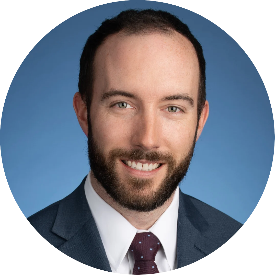 Smiling man with short dark hair and beard wearing a navy suit, white shirt, and maroon tie with white dots, posed in front of a solid blue background.