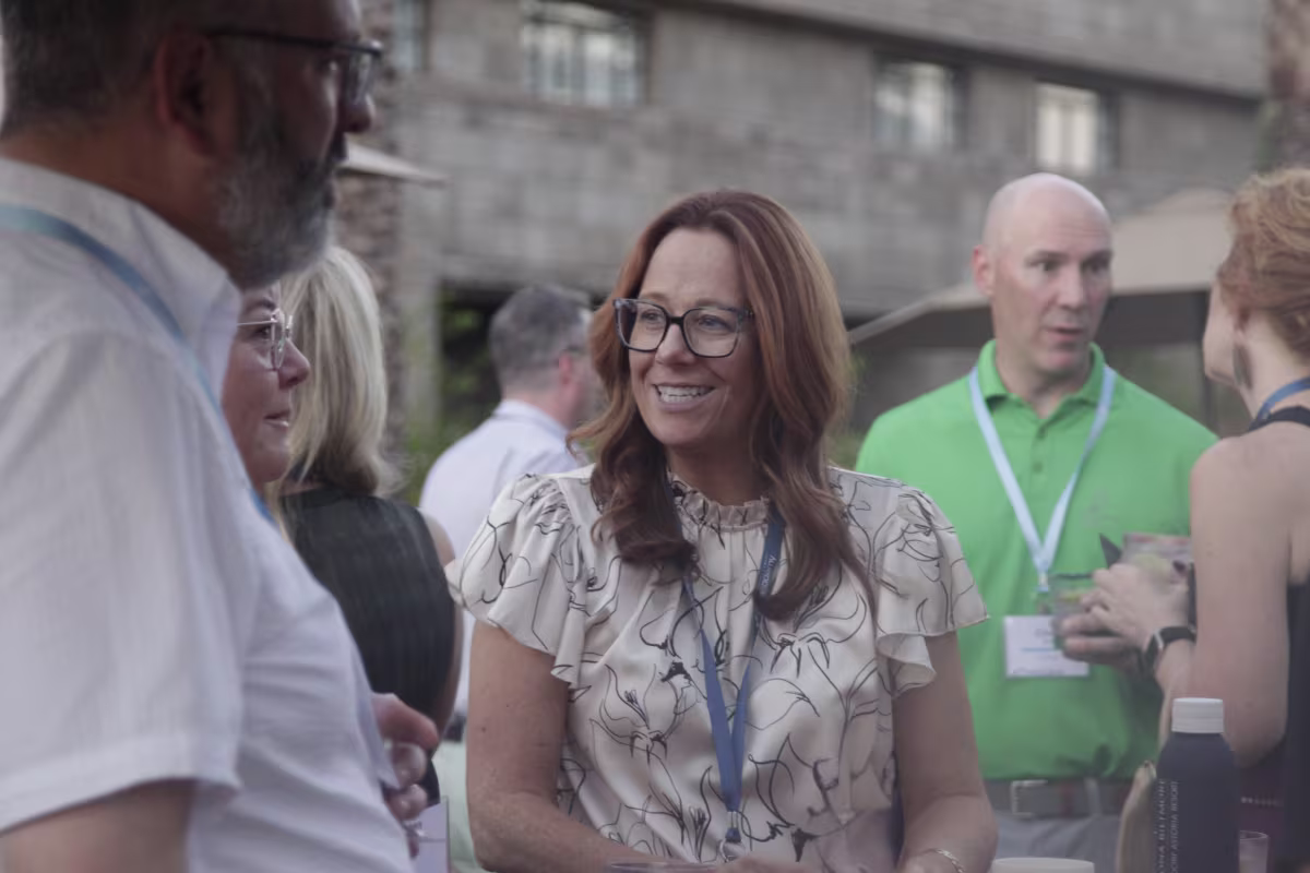 A woman with long auburn hair and glasses, wearing a white blouse with a black floral pattern, smiles while conversing at an outdoor networking event. She holds a canned beverage, and other attendees around her hold drinks and chat in the background.
