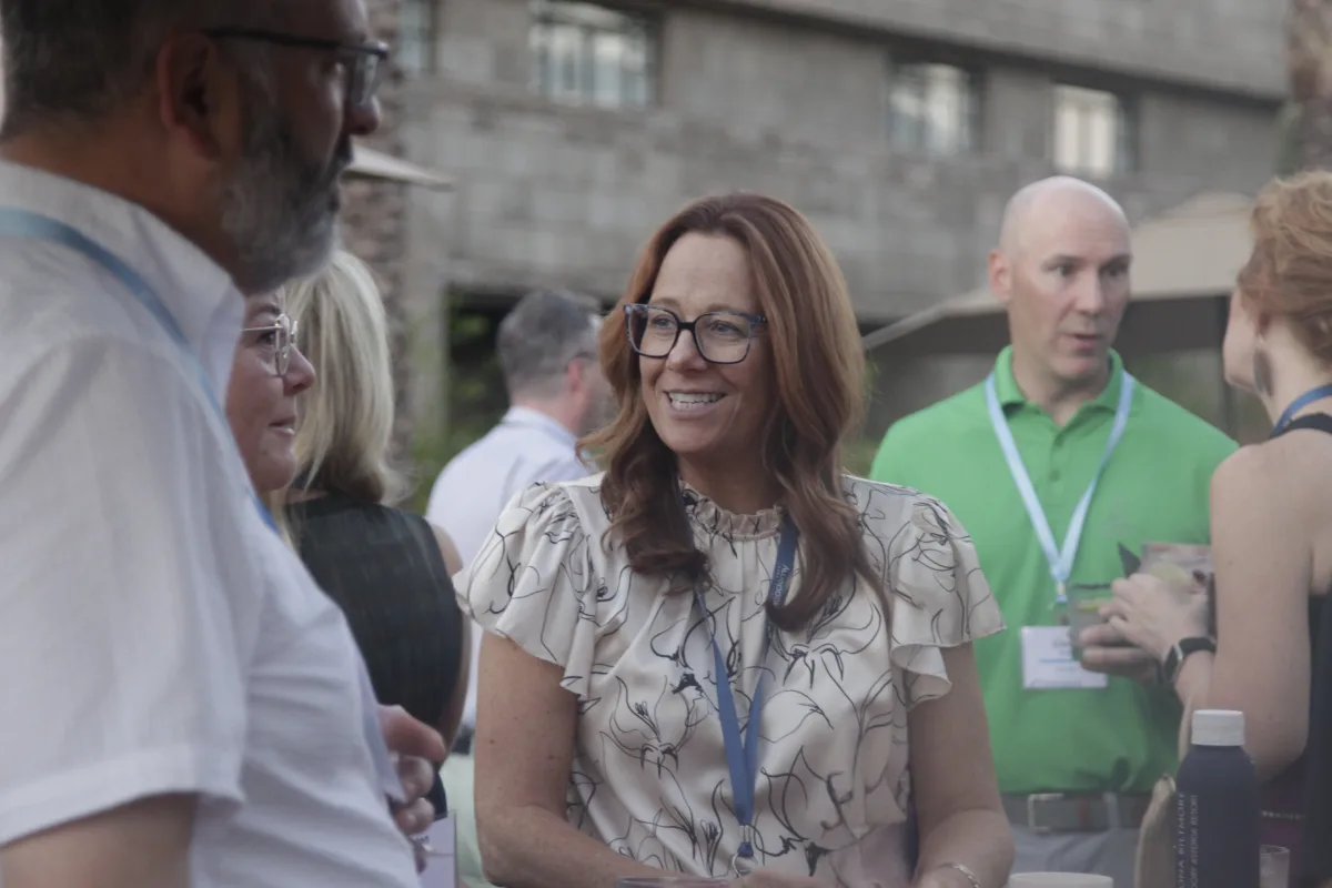 A woman with long auburn hair and glasses, wearing a white blouse with a black floral pattern, smiles while conversing at an outdoor networking event. She holds a canned beverage, and other attendees around her hold drinks and chat in the background.
