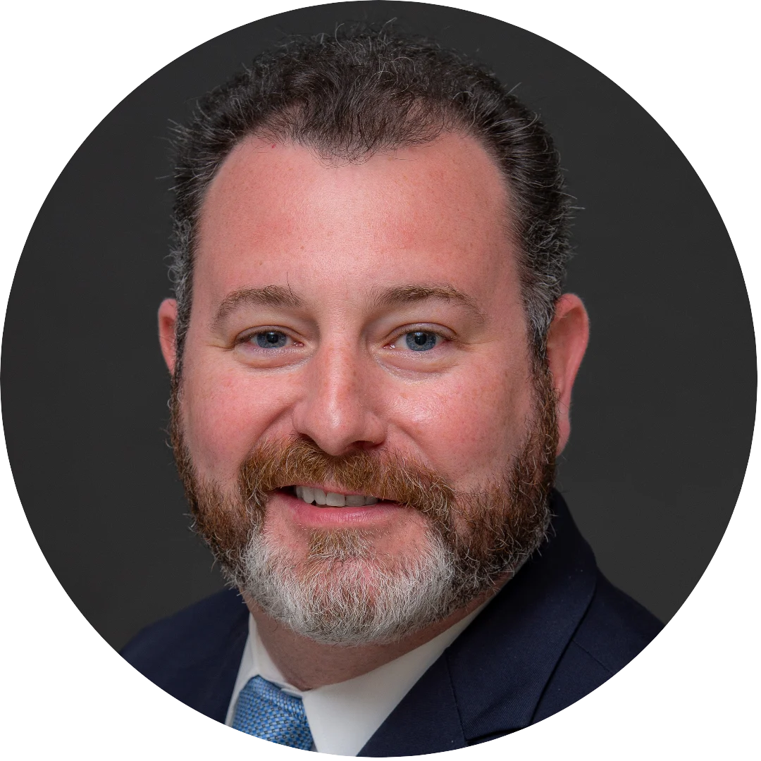 Smiling man with a short curly salt-and-pepper beard and hair, wearing a navy suit, white shirt, and light blue tie, posed against a dark gray background.