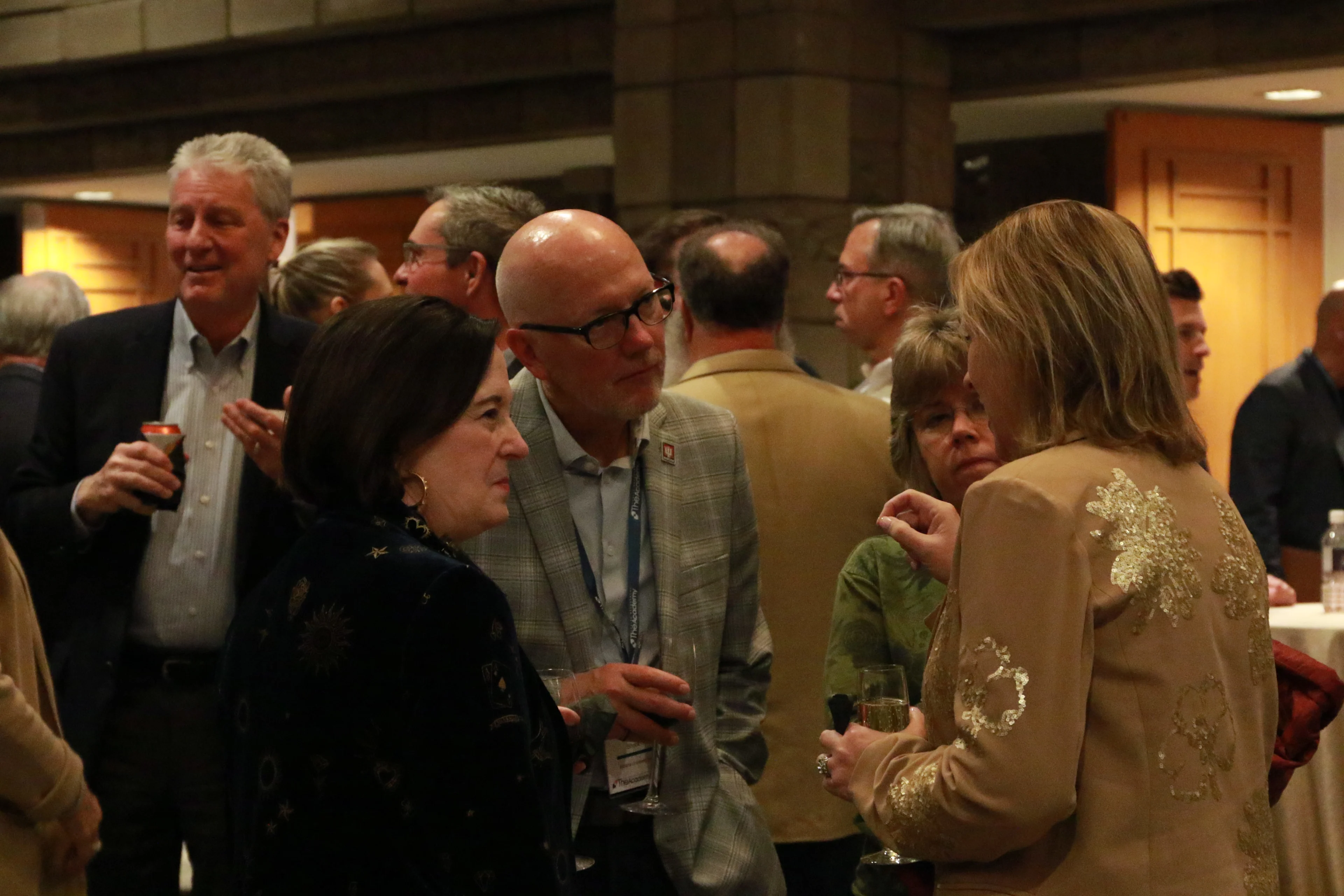 A group of people engage in conversation at a networking event. In the foreground, a man in glasses and a gray plaid blazer, a woman in a dark patterned jacket, and a woman in a beige jacket with gold floral embroidery speak while holding drinks. The background shows other attendees mingling in a warmly lit room.
