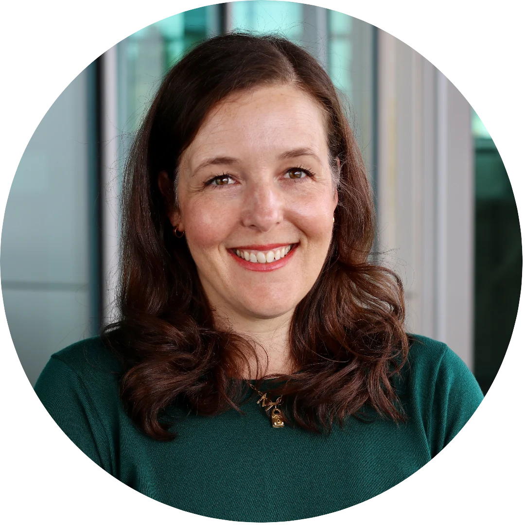Smiling woman with long wavy brown hair wearing a dark green top and gold necklace, posed indoors in front of a glass-panel background.