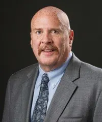 Professional headshot of a middle-aged man with a bald head, mustache, and light complexion, wearing a gray suit, patterned tie, and light blue shirt, smiling against a dark background.