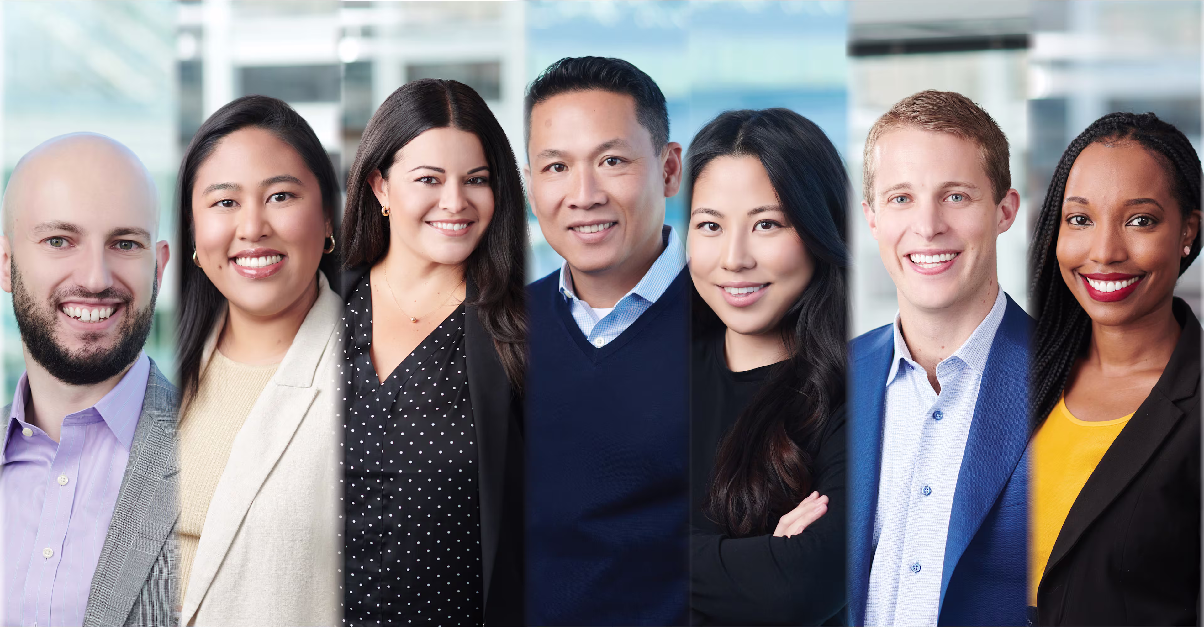 Group portrait of seven professionally dressed individuals standing side by side in a modern office environment, all smiling at the camera.