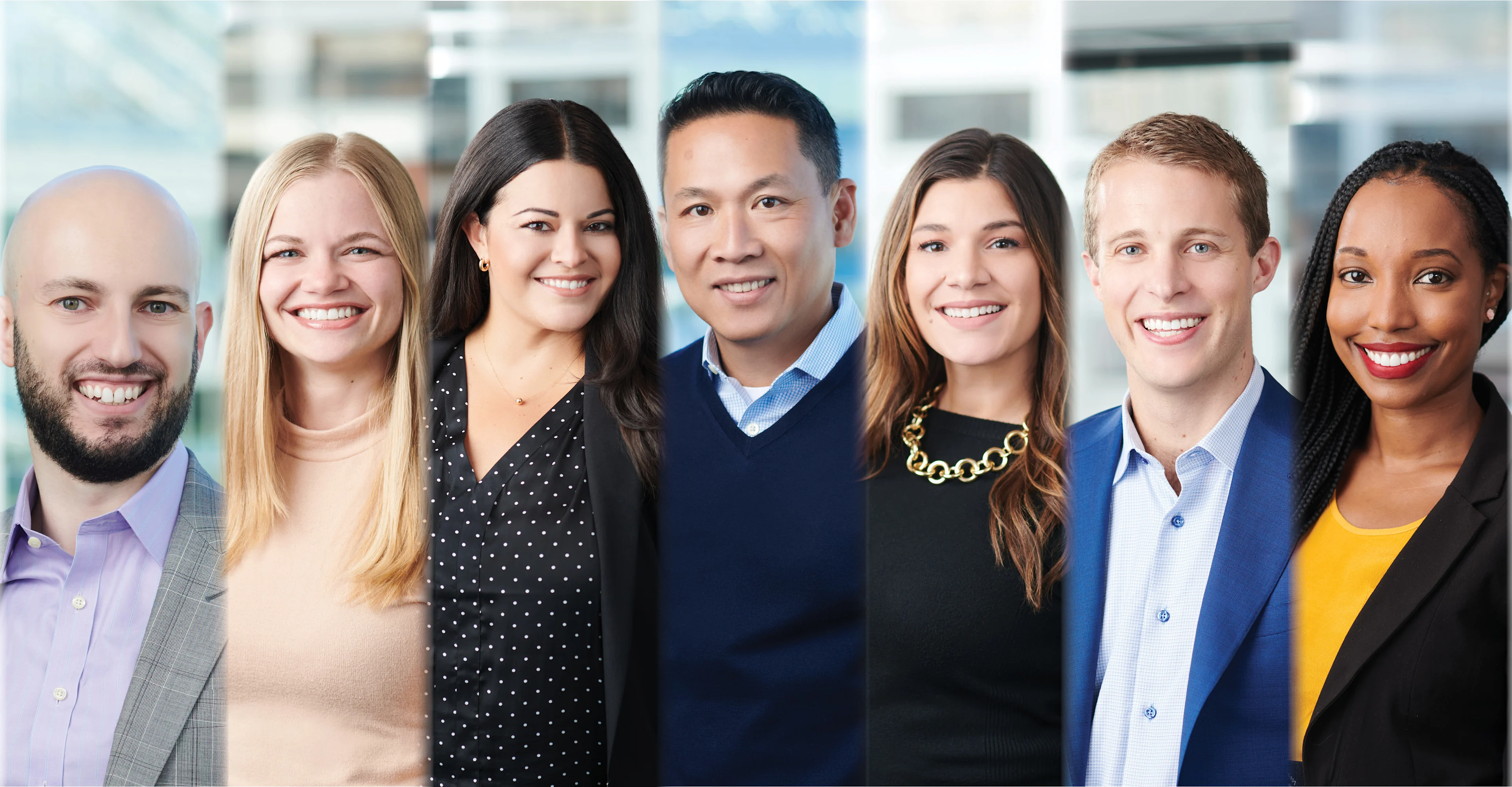 Group portrait of seven professionally dressed individuals standing side by side in a modern office environment, all smiling at the camera.