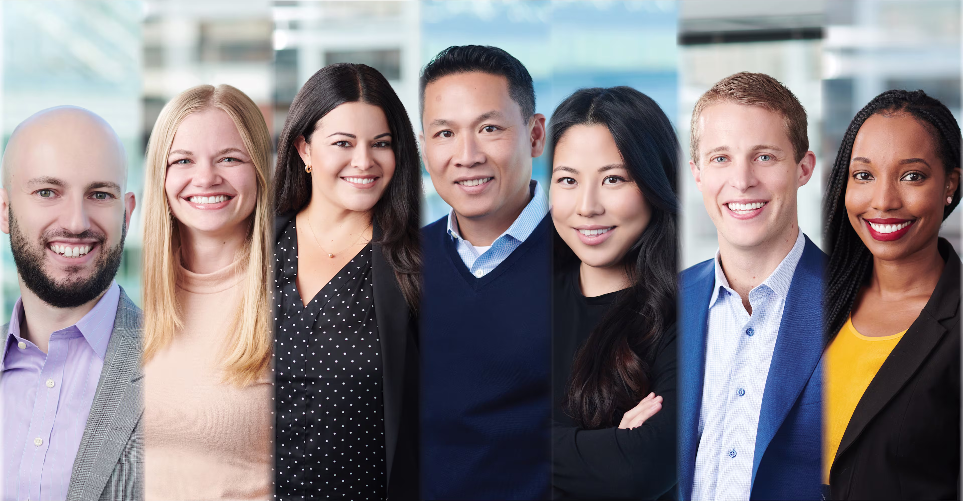 Group portrait of seven professionally dressed individuals standing side by side in a modern office environment, all smiling at the camera.