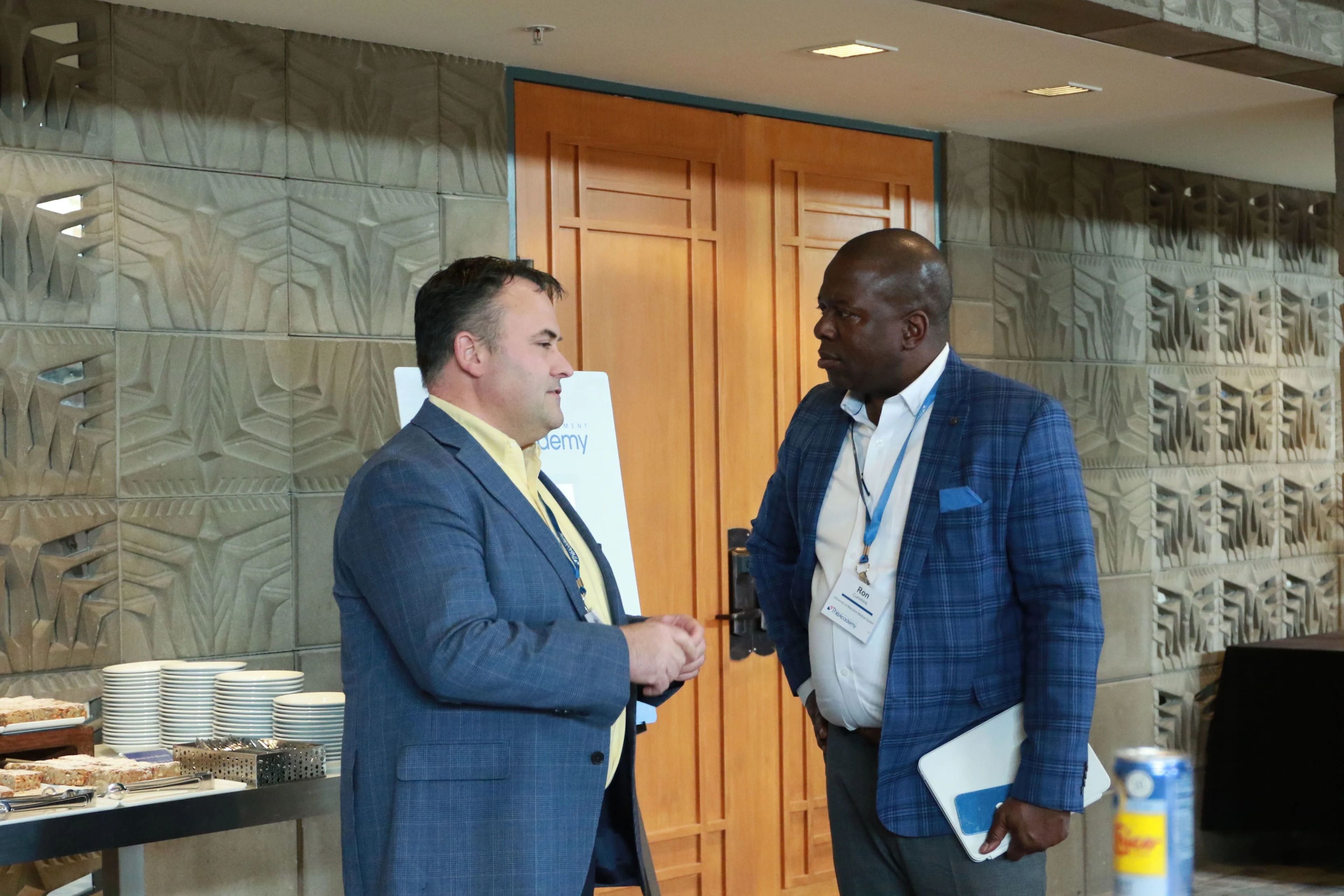 Two men in business attire stand and converse near a refreshment table at a professional event, with one holding a folder and the other gesturing mid-discussion.

