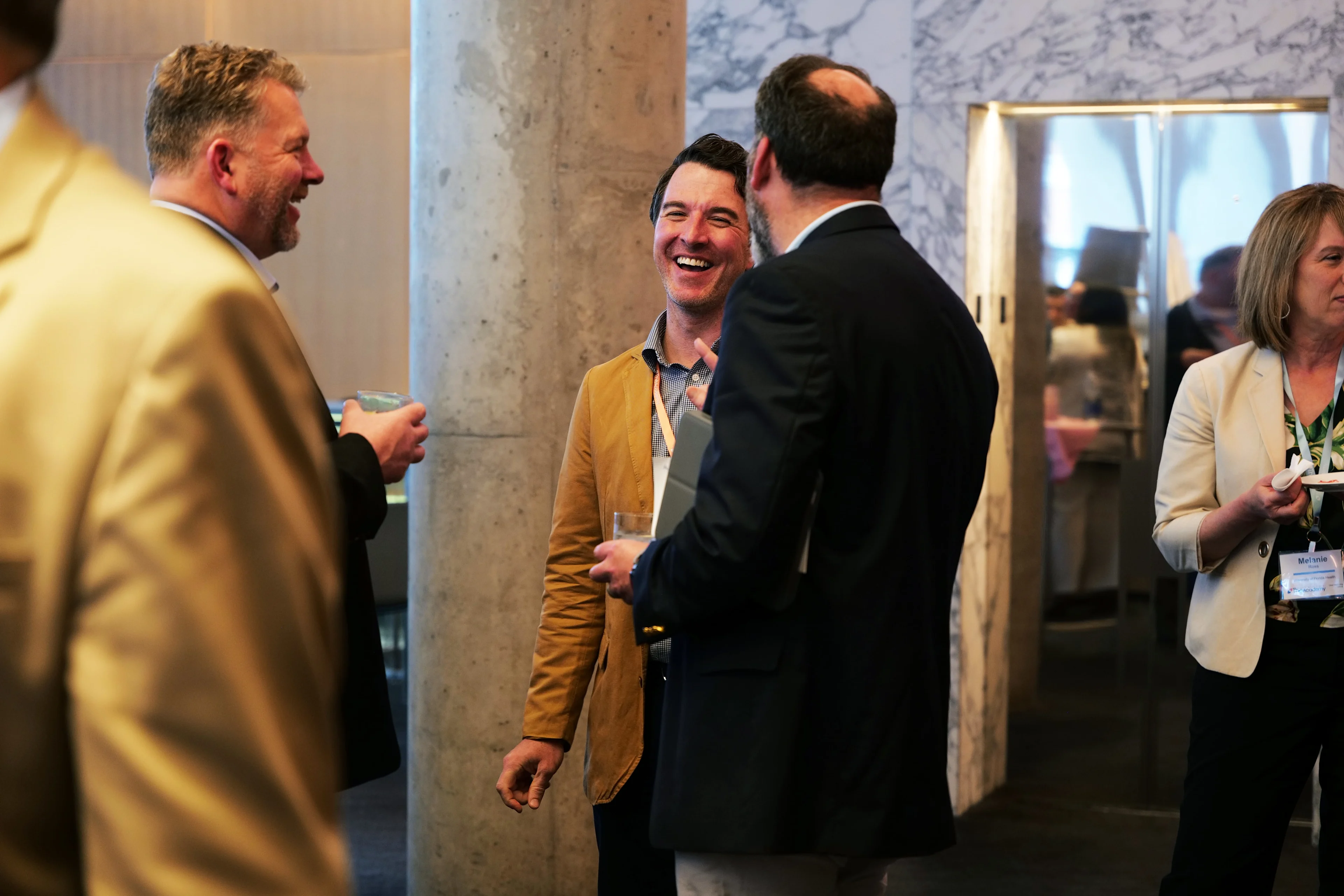 A group of professionally dressed attendees engage in casual conversation at an executive event. One man in a tan blazer is smiling and holding a drink while speaking with others. The setting includes a marble wall, a concrete pillar, and warm lighting, creating a relaxed and professional networking atmosphere.

