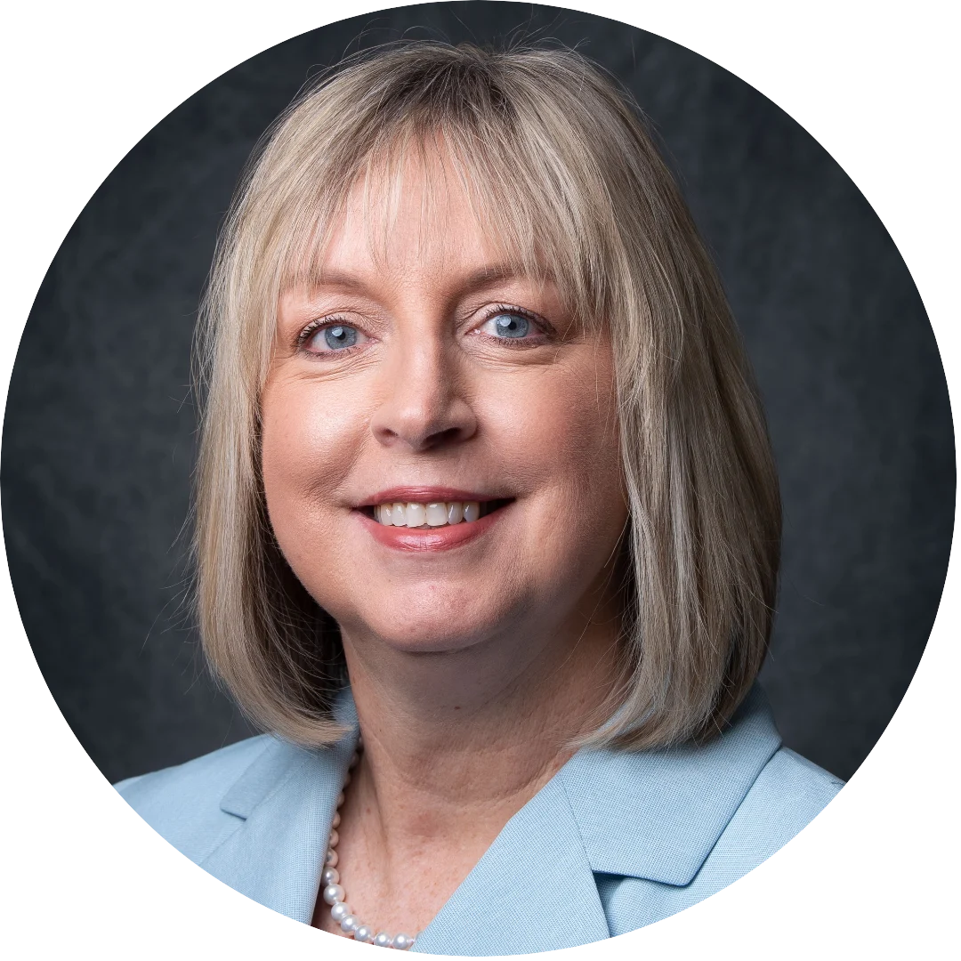 Woman with straight blonde hair wearing a light blue blazer and pearl necklace, smiling for a formal headshot against a dark textured background.
