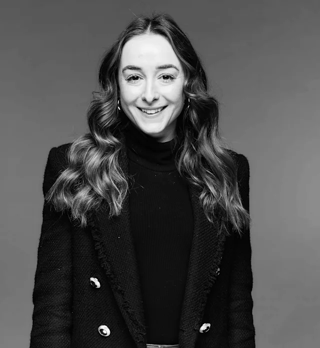 A woman with long wavy hair smiles in a black and white portrait, wearing a dark double-breasted blazer and a turtleneck against a plain backdrop.








