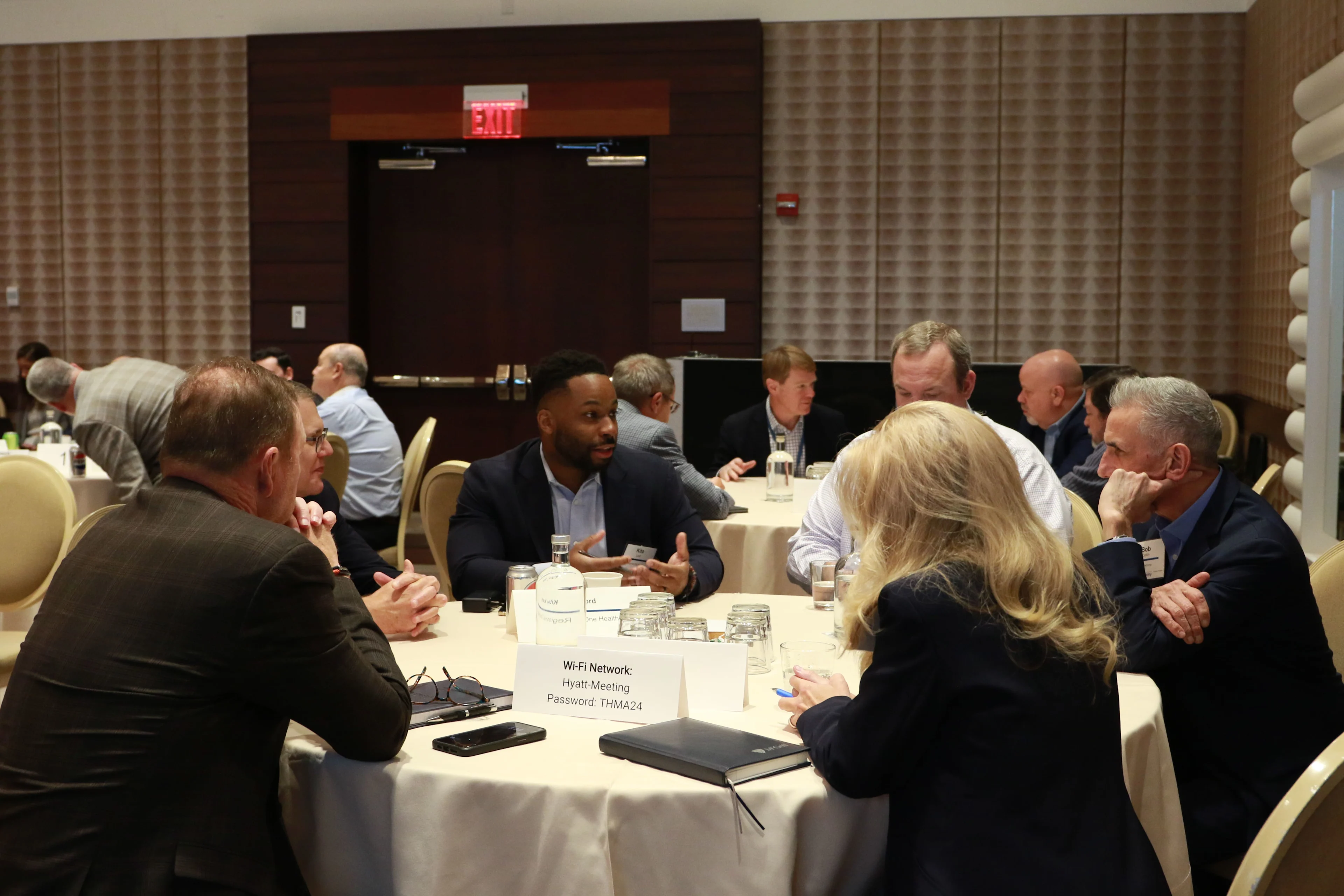 A group of professionals sits around a roundtable engaged in conversation during a formal discussion session. The setting appears to be a conference or leadership forum, with name cards, notebooks, and glasses of water on the table. The background includes other participants seated at similar tables in a well-lit meeting room.

