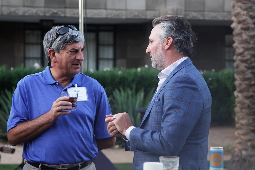 Two men converse during an outdoor networking event, one holding a drink and the other gesturing while speaking. They are dressed in business casual and formal attire.