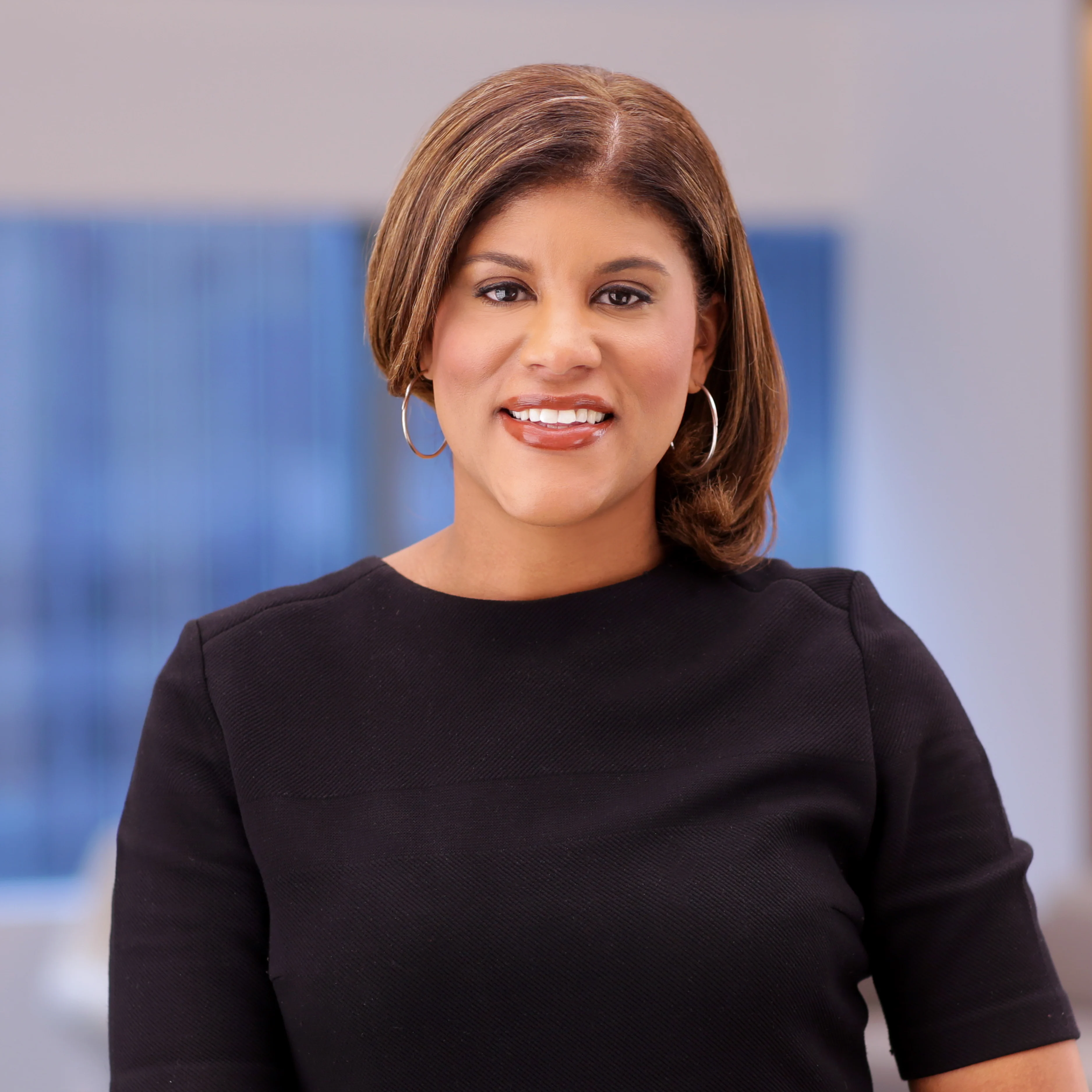 A woman with shoulder-length brown hair and hoop earrings smiles while wearing a black dress. She stands in a softly blurred indoor setting with cool-toned background lighting.