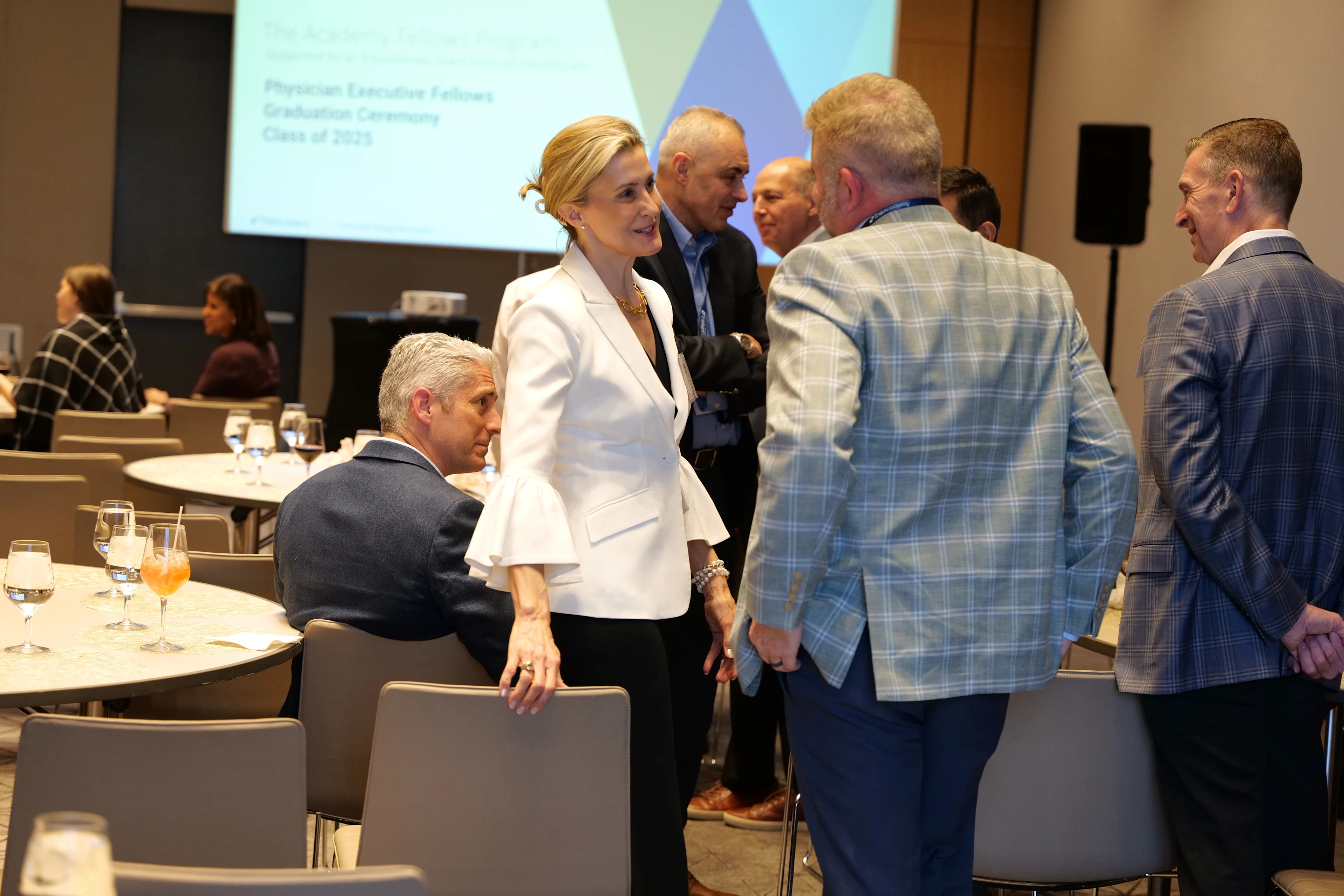 A woman in a white blouse is speaking with a man in a blue plaid jacket during a networking session at the Physician Executive Fellows Graduation Ceremony, with other attendees conversing in the background.
