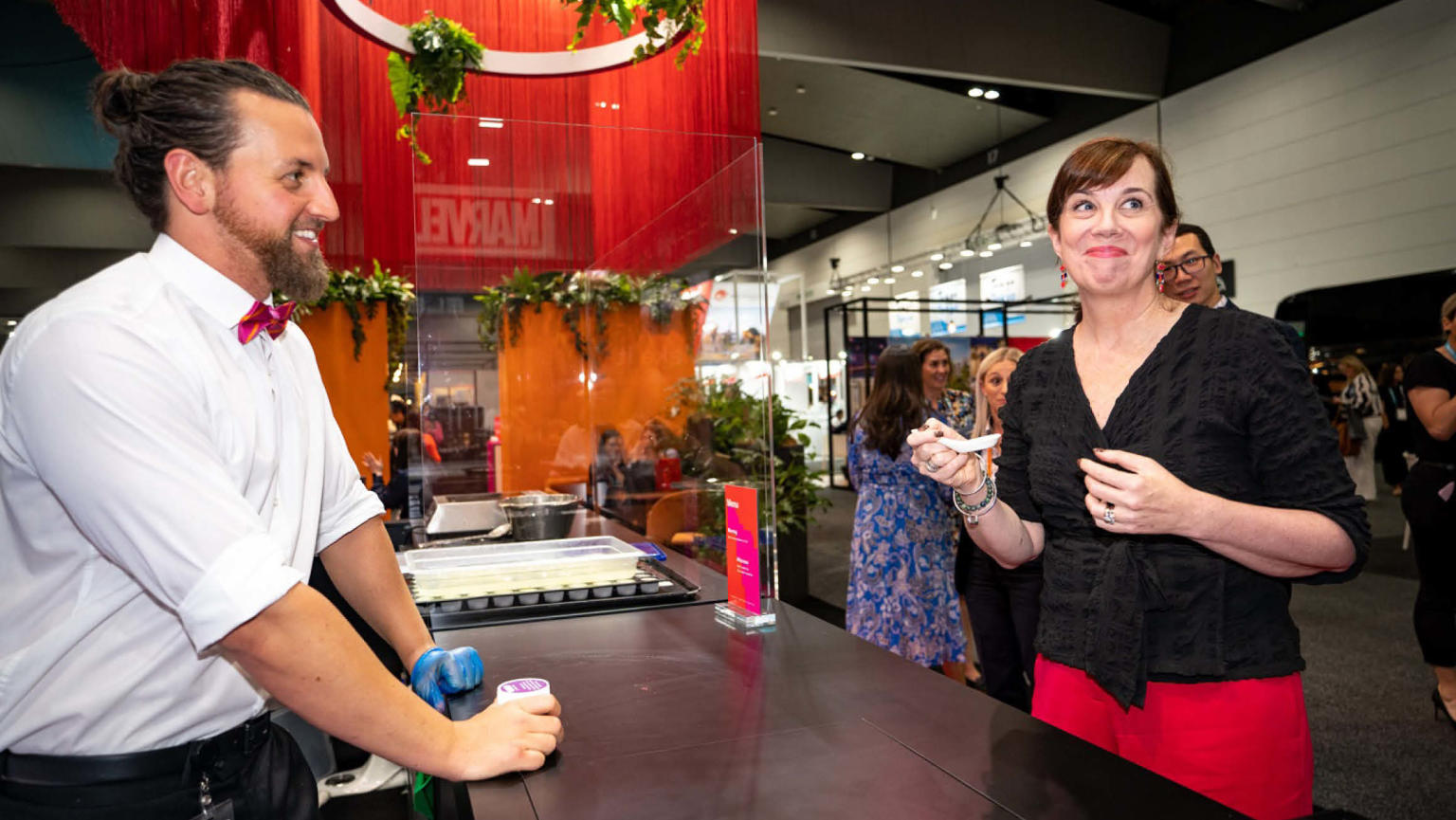 A gentleman wearing a white shirt and a stylish red bow tie stands cheerfully behind a bench, while a woman dressed in a black shirt and pink pants smiles while holding a spoon and savouring a delectable treat.