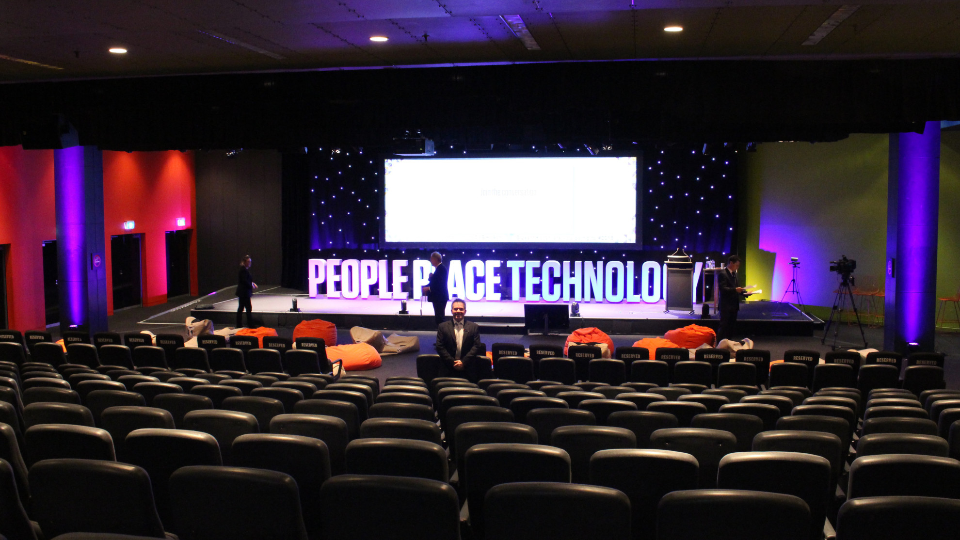 A man stand facing the camera amongst rows of black seats. A stage with large lit up letters saying People Place Technology under a large white screen are in the background. 