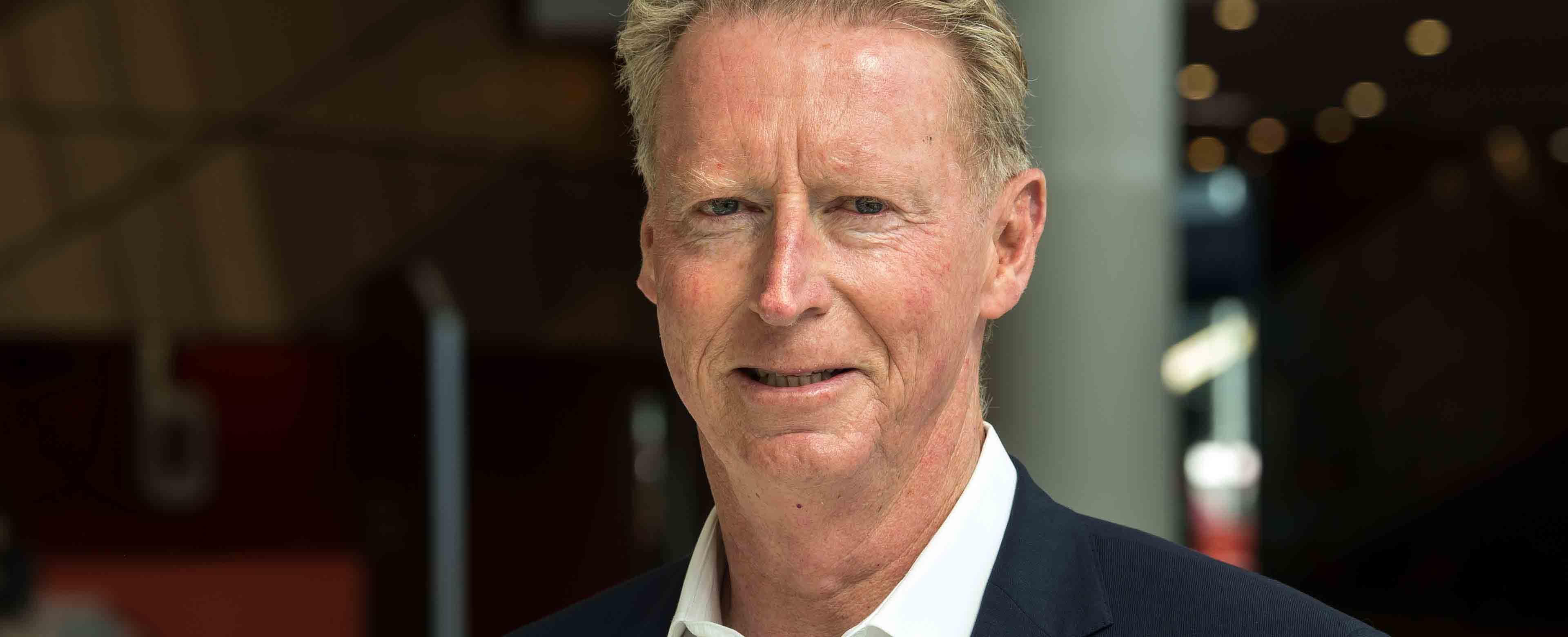 Headshot of former Melbourne Convention and Exhibition Centre Chief Executive, Peter King, wearing a suit and smiling at the camera. The convention centre atrium is visible in the background.